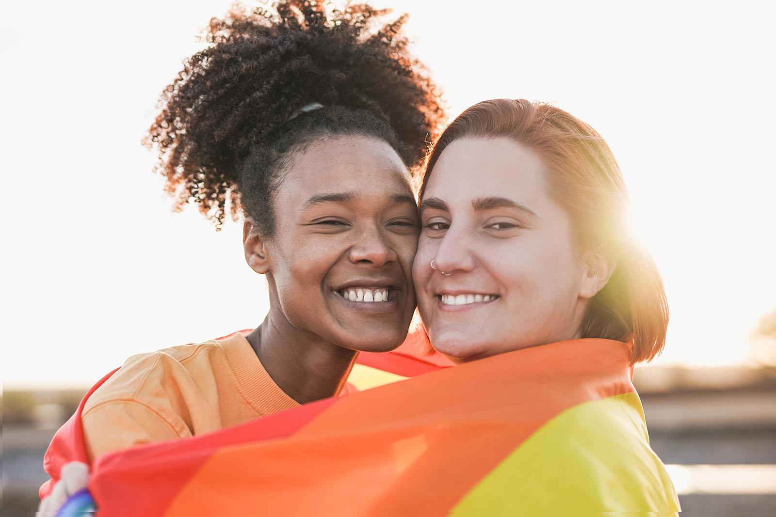A multiracial couple hug with a pride flag wrapped around them outside