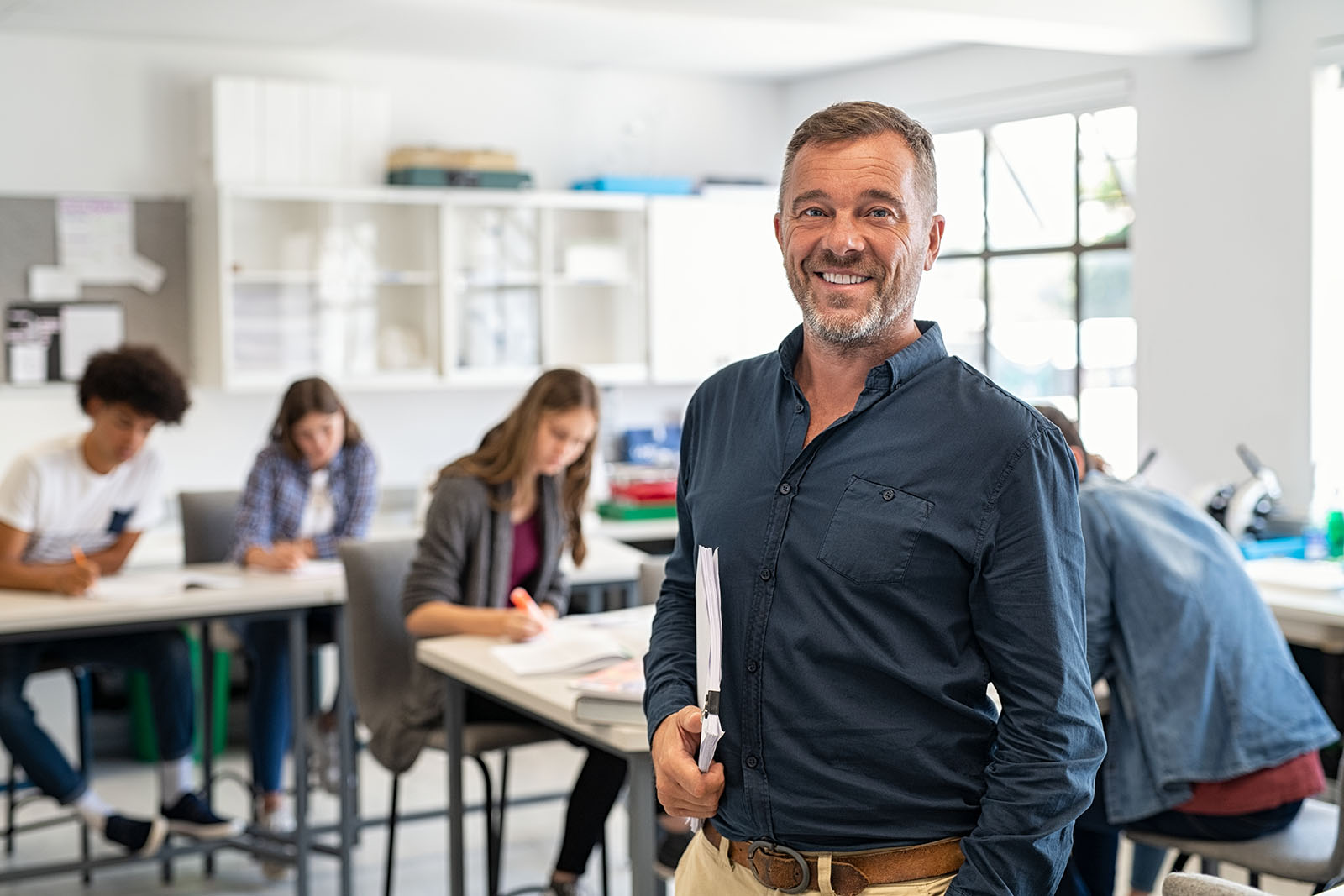 A man in a classroom smiling and holding papers