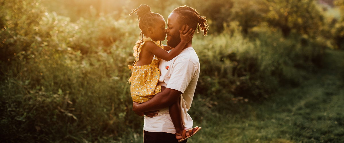 A father holds his daughter in his arms and their noses touch during a sunset outside