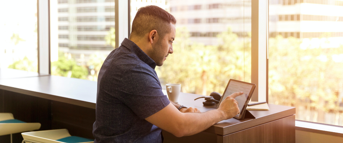 A man uses a tablet while sitting at a desk with a coffee