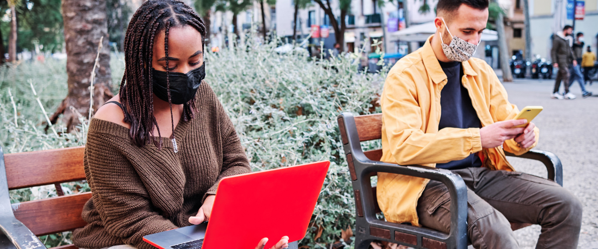 People sit outside with electronic devices in a public park wearing masks
